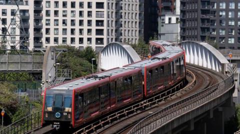 A Docklands Light Railway train crosses a bridge. There are residential blocks in the background.