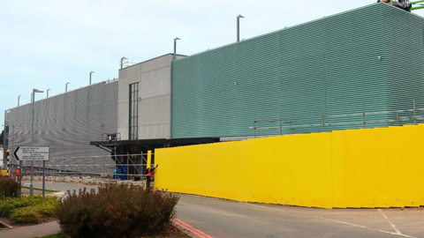 A large multi-storey car park covered in grey and green cladding. A yellow site fence is partially surrounding the building revealing scaffolding behind it. 