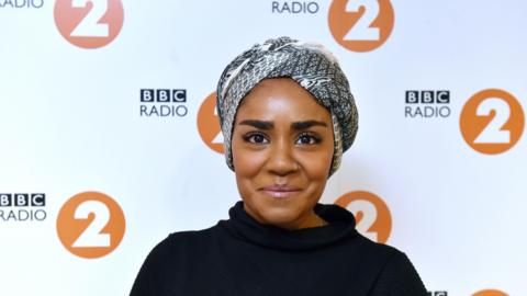 A woman is stood in front of a white background with BBC Radio 2 logos spaced out across it. She is wearing a black and white hijab and a black top.