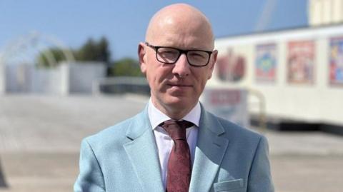 Patrick Hurley has a bald head, dark spectacles, a white shirt, maroon tie and sky blue jacket,