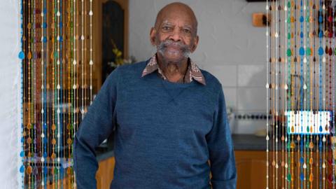 Alford Gardner, an elderly gentleman wearing a blue jumper and bushy stubble, smiles into the camera while standing in a kitchen.