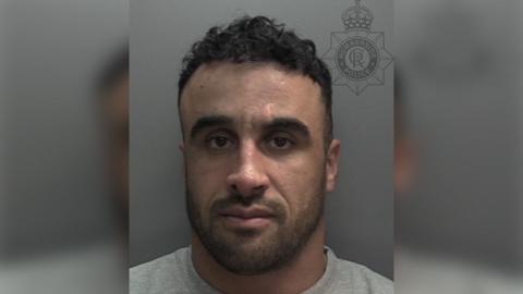 Police custody photograph of Robert Kel. He has short black hair and a beard and is wearing a grey T-shirt