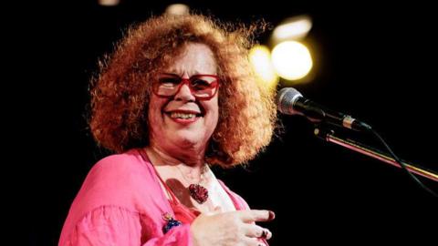 A woman, Sarah Jane Morris, wearing a pink top and standing on a stage with a microphone.