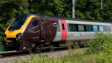 A Crosscountry train with the cross part of the logo written in red and the country part of the logo written in white. It is all in lower case and visible on the left hand side of the train, which is in shot. The doors of the train are red and the front-facing end of the train is painted yellow. The rest of the train visible has a burgundy section and a grey section. 
Behind the train trees are visible, with a lamp post by the side of the train track. 