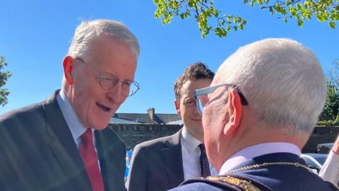 Shows a man on the left with grey hair and glasses. He is wearing a dark jacket, light blue shirt and red tie. He is standing next to a younger man wearing a dark jacket, white shit and dark tie and a man with his back to the camera wearing a mayoral chain around his neck. He has grey hair and is wearing glasses. 