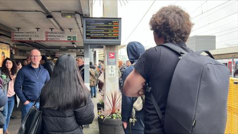 People standing on a busy platform at Colchester railway station. Most people in the picture have their backs to the camera and are looking at a travel information screen, which shows all journeys being delayed. It is a grey day and most people are wearing coats or jumpers.