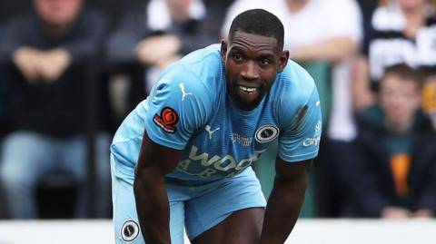 Lee Ndlovu crouching down with a slight grimace on his face mid-match. He is wearing light blue Boreham Wood kit.