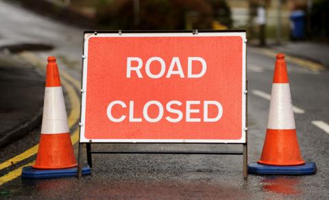 A red 'road closed' sign with white writing is pictured on a road with two traffic cones on either side. Behind and beside it, you can see white and yellow road markings.