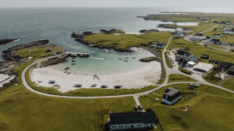 An aerial view of a small sandy bay and harbour in Tiree. The low-lying, grassy landscape around the bay is dotted with houses and other buildings.