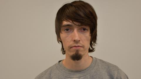 A custody photo of a man with brown hair swept across his foreheard. He is against a plain grey background, and is wearing a light grey sweatshirt. He has a short goatee beard.