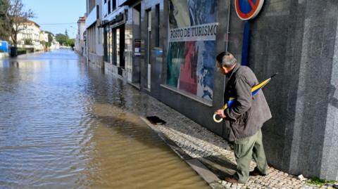 A man holding an umbrella stands on a pavement looking down a flooded street.