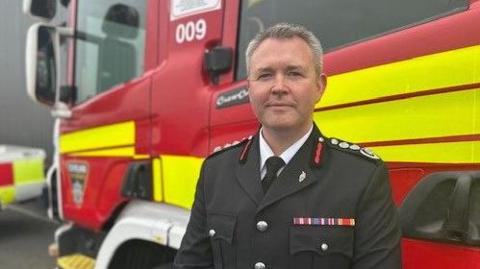 Peter Rickard, chief fire officer at Cleveland Fire Brigade. He has short grey hair and is wearing a black uniform with a white shirt and black tie. He is standing in front of a fire engine and looking directly into the camera.