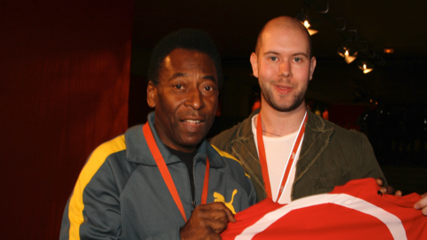 Pele and Rob are standing together holding a red football short that is slight obscured. Pele is in a navy and yellow striped tracksuit and Rob is wearing a grey jacket and white t-shirt. He has a shaved head and short beard.