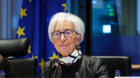 Head of the European Central Bank, Christine Lagarde, sitting wearing a black leather jacket and a black and white scarf around her neck, with the EU flag behind her