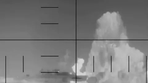 Screengrab from a video showing a black and white image of a ship on fire, with a spout of water rising from the foreground and crosshairs in front