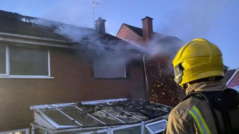 A firefighter in full gear looks at a damaged house. It has a hole in the roof and a window blown out. Debris can be seen on top of the conservatory. 