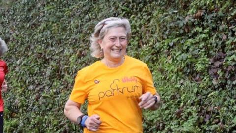 Yvonne Turner running and smiling. She is wearing a yellow top with the word 'Parkrun' on the front.