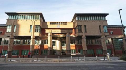 Sheffield Crown Court from the outside, a large four-storey building with pillars at the front. Four trees lines the street in front of the building and there is a line of bollards in front of a road that runs outside the court.