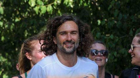 Celebrity fitness coach Joe Wicks running during a Parkrun event. There are other runners smiling and chatting around him, and he is smiling straight at the camera.