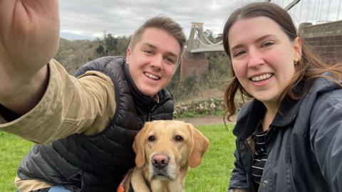 Adam Drysdale and his partner, Elle Manners, pictured by the Clifton Suspension Bridge. Adam has short brown hair and is clean shaven. Elle has long dark hair. Both of them are crouched down in a selfie with a blonde labrador. 