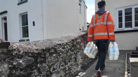A person in hi-vis carries a number of plastic water bottles to a house.