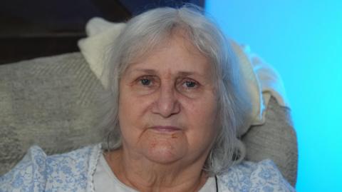 A woman in her seventies with grey bob-length hair sits in a light-grey easy chair with he head resting on a white blanket. She is wearing a blue and white dress. The background is bright blue.