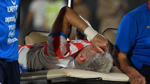 Julio Enciso holds a hand over his face while being stretchered off