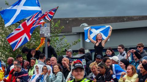 A large group of people are gathered at a barrier. Union and Saltire flags fly above the group. There is a woman, standing on an unseen wall, with arms raised and holding a Saltire.