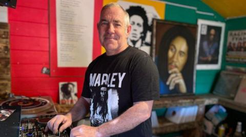 Robert Flake wearing a black t-shirt with a picture of Bob Marley and his name printed on it, standing in his shed that is filled with his records, a record player and posters of Marley