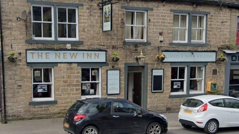 Street view image of the New Inn. It is a double fronted stone building with pale blue and gold signage and white framed windows. There is a small black car and a small white car parked outside