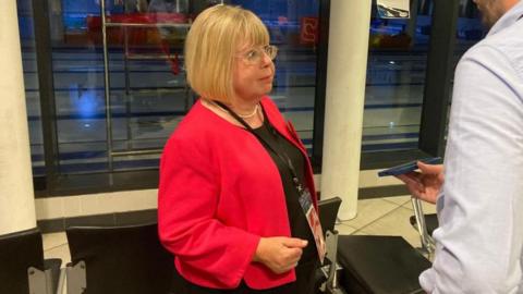 Liz Twist being interviewed by a reporter. She has blonde hair and is wearing glasses. She is also wearing a red blazer and a lanyard. A reporter to her left is holding a phone out to record their conversation.