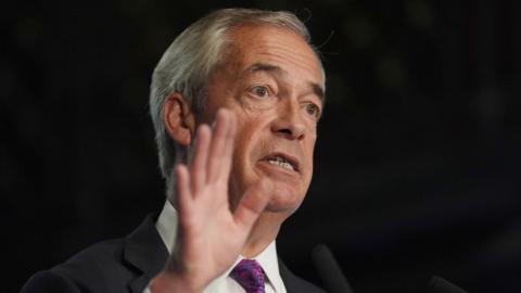 Nigel Farage wearing a suit and tie with his hand in the air. He has white hair 
