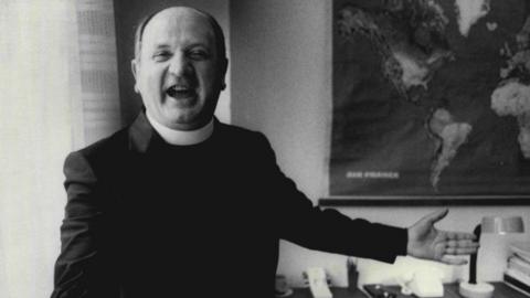 A black and white image of Bishop Eamonn Casey, wearing a black suit and smiling. There is a map of the world on the wall behind him. 