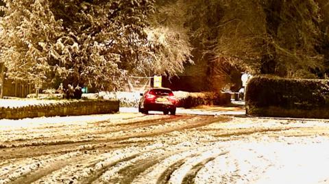 A car with its brake light showing drives on a snowy road. There are several vehicle tracks in the snow with snow dusting hedges and walls on either side