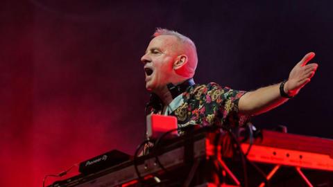 Fatboy Slim performing onstage during a concert. He is standing in front of a DJ set and is holding his arms up.