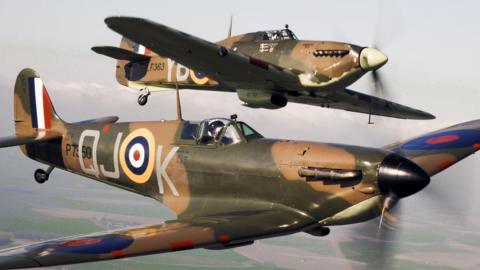 Two World War Two-era fighter planes, a Spitfire and a Hurricane, fly side by side over countryside against a grey sky. The planes are painted in a green and brown camouflage scheme and have RAF red and blue roundels on the wings and red, white, blue and yellow roundels on the fuselage, along with grey and black identification numbers. The tails have red, white and blue RAF vertical stripes.