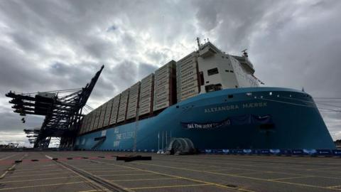 A large container ship at the port of Felixstowe. It is blue and has white shipping containers stacked up on top of it.