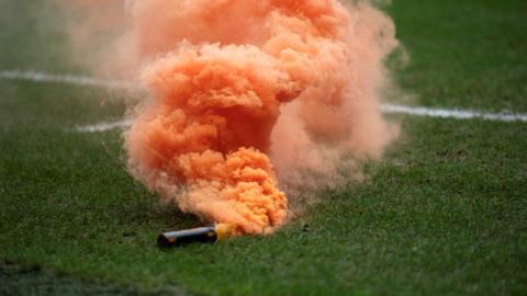 A pyrotechnic omitting orange smoke onto a football pitch.