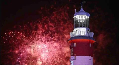 A red firework explodes against a dark sky and produces a sparkling backdrop to a lighthouse which has red and white stripes