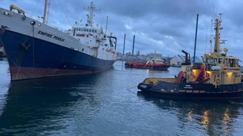 A cloudy image of a shipyard with a vessel called the Empire Persia in the foreground and two tugs around her 