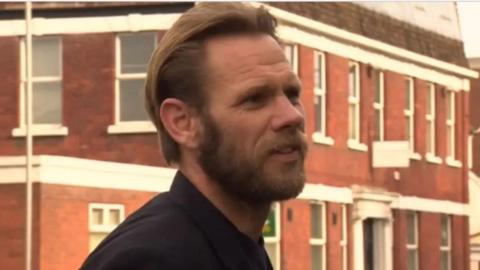 A head and shoulders shot of Robert Jenner wearing a dark shirt with swept back ginger hair and a beard on a street with houses behind him