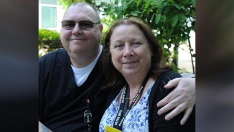 John Ellington is wearing a dark V-necked sweater over a white T-shirt, and tinted glasses. He has his left arm round Sandy Ellington, who is wearing a dark cardigan over a blue and white patterned blouse. She is wearing a lanyard with a name badge on it. Both are smiling. They are outside with a hedge in the background.