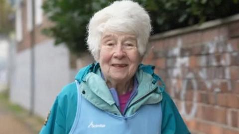 Barbara Thackray with white hair wearing her running gear including a green jacket standing in front of a brick wall with graffiti on it. She is smiling.
