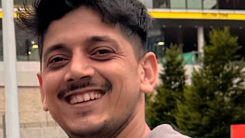 A man with short black hair and a short stubbly moustache and beard wears a grey zipped hoodie and black t-shirt while smiling for a photo