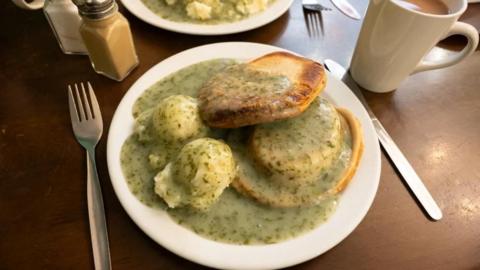 A plate of pie 'n' mash sitting next to a silver knife and fork, a white cup and salt and pepper pots. The pie 'n' mash is covered in a parsley sauce.