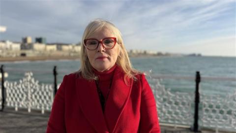 A woman wearing a red coat and red glasses. She is stood on a pier with the sea and seafront in the background.