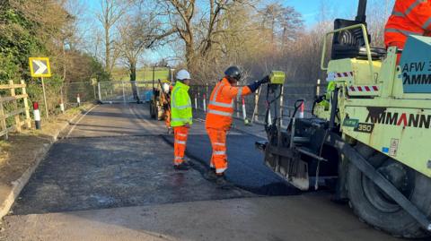 The A361. There are men in hi-vis jackets guiding a large tractor like machine that appears to be laying down tarmac.