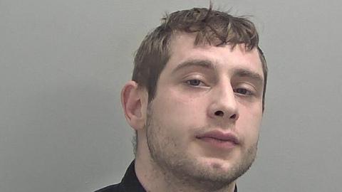 A mugshot of Oliver Beck. He has brown hair and slight facial hair and is wearing a black top.