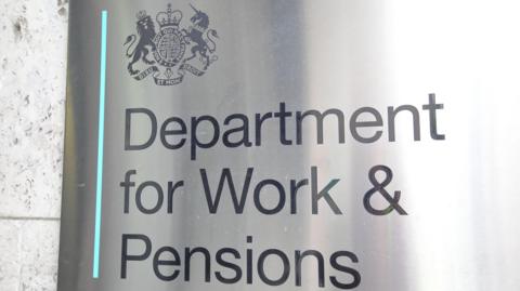 Close-up of a grey metal sign which says "Department for Work and Pensions". The government's logo containing a unicorn and a lion is above it in the top left corner. 