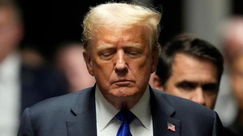 Donald Trump stands with his eyes half closed. He is wearing a suit with a blue tie and American flag pinned to his collar